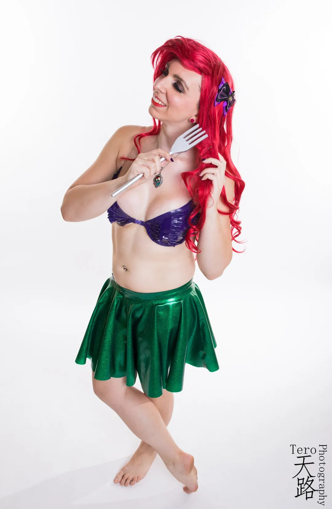 Jerikandra as Ariel from The Little Mermaid with red hair, purple seashell top and green skirt, holding a fork, on a white backdrop.