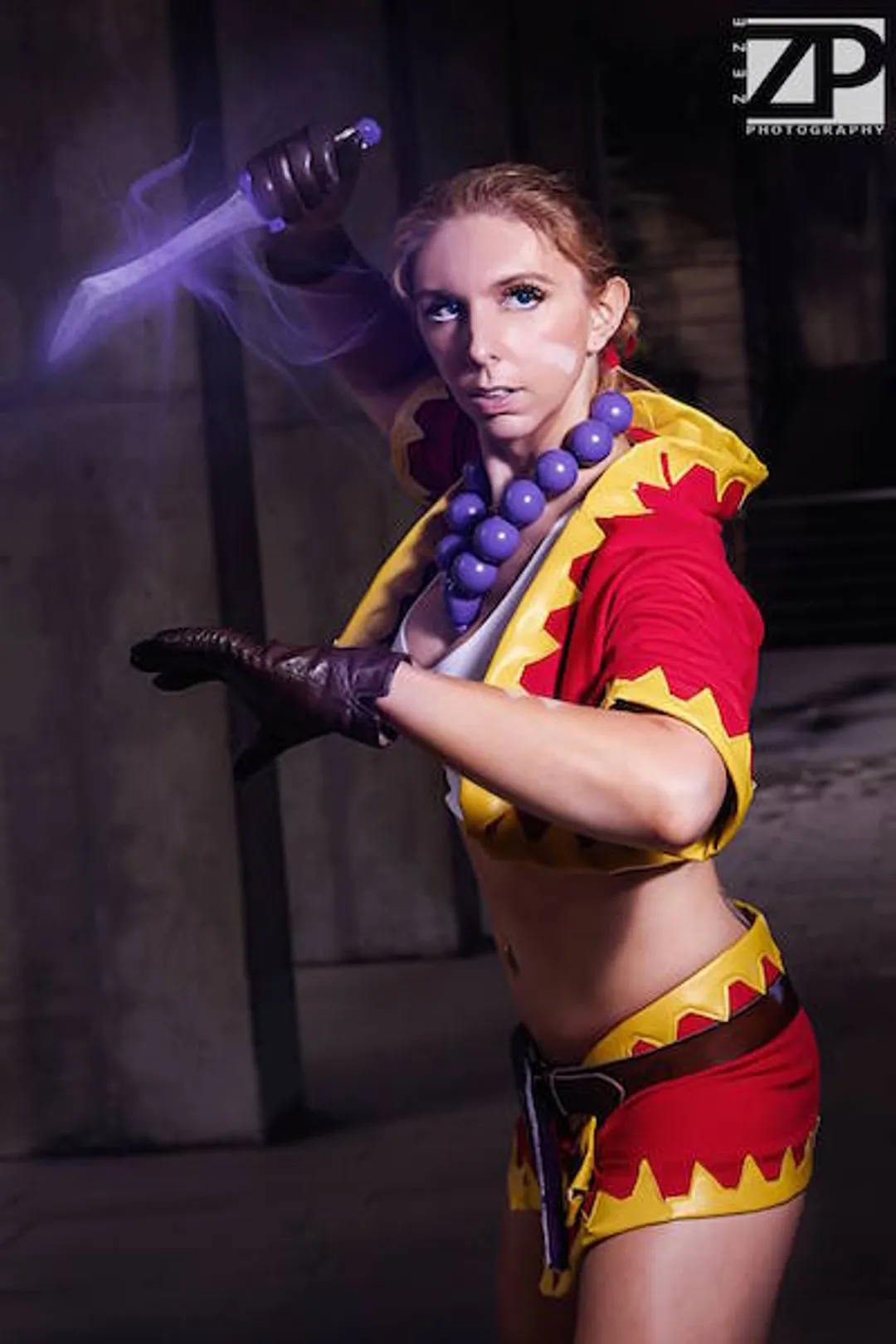 Jerikandra as Kid from Chrono Cross in a red and yellow zigzag-trimmed outfit with purple bead necklace, holding a glowing dagger overhead.