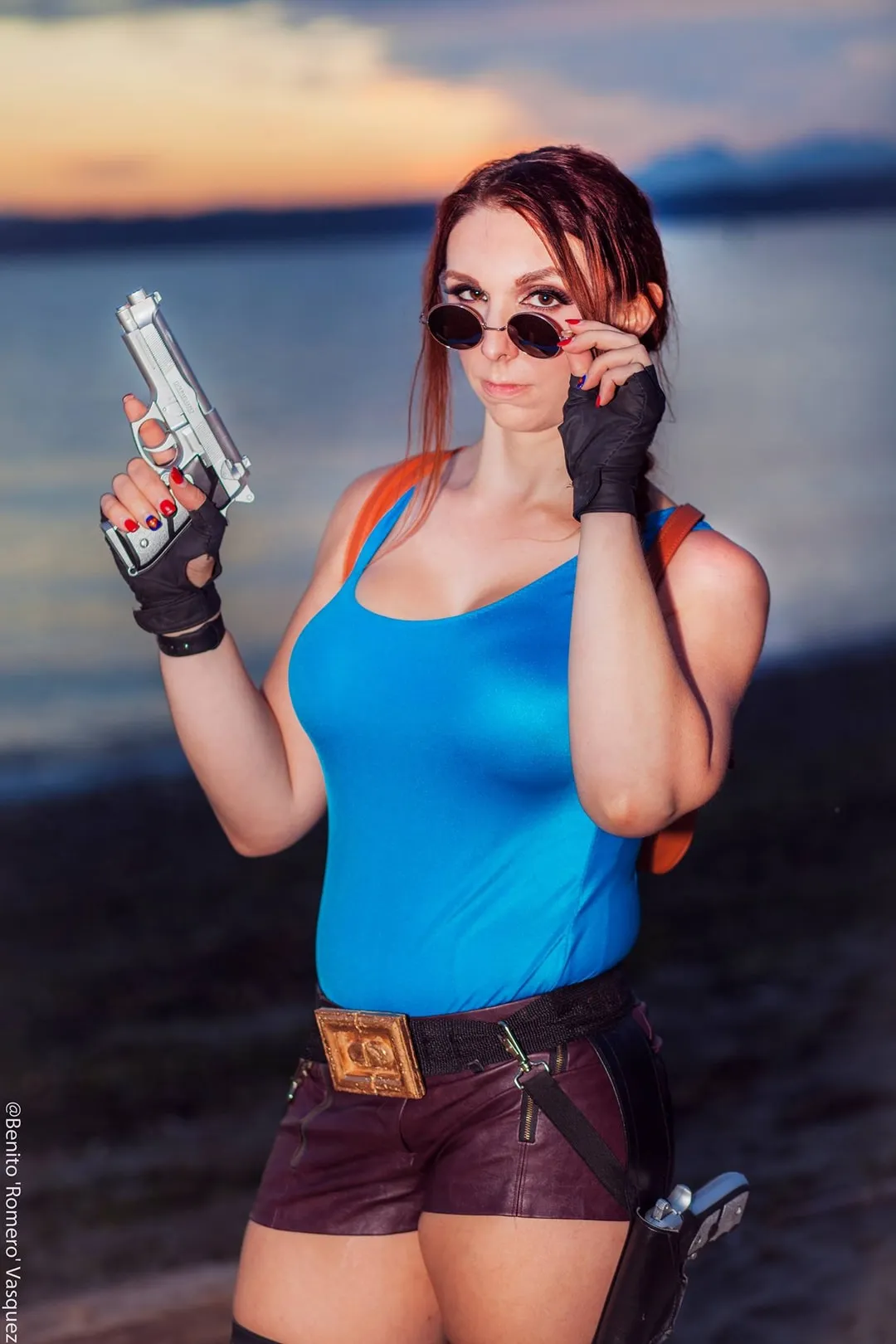Jerikandra as Lara Croft in her classic blue tank top and brown shorts with a pistol and sunglasses, at a sunset beach.