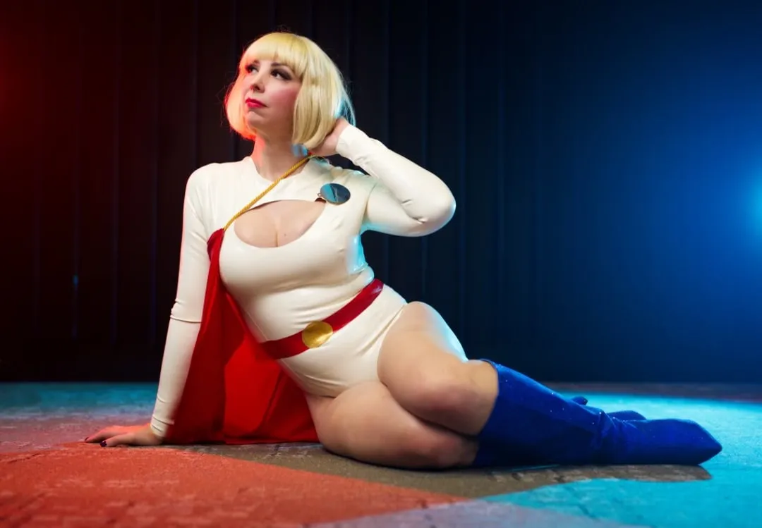 Jerikandra as Power Girl in a white leotard with red cape and blue boots, posed reclining against a blue studio backdrop.