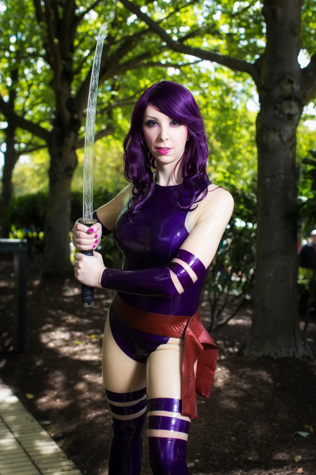 Jerikandra as Psylocke in a purple latex suit with red sash and purple hair, holding a sword under green trees.