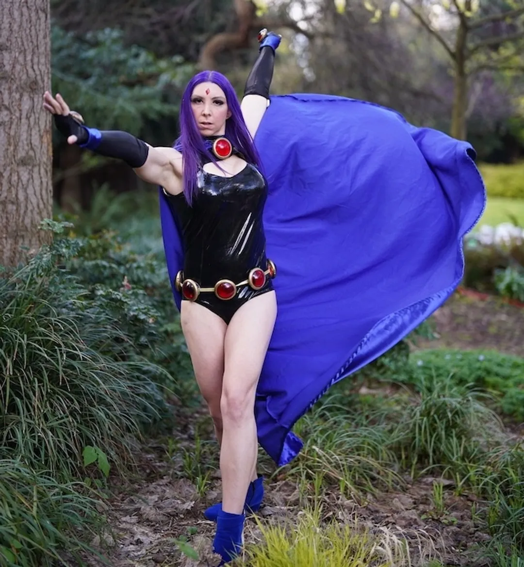 Jerikandra as Raven from Teen Titans in a black latex leotard and flowing blue cape, one arm raised, outdoors among trees.
