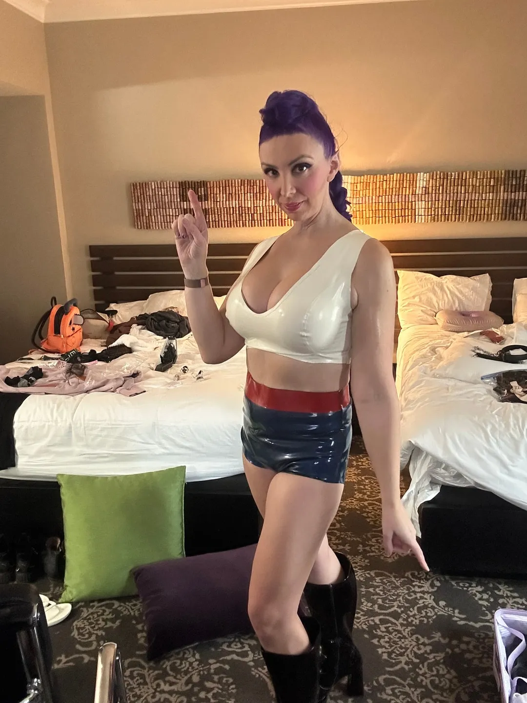 Jerikandra as Rumi from KPop Demon Hunters with a purple braid, white latex top and striped shorts, posing in a room.