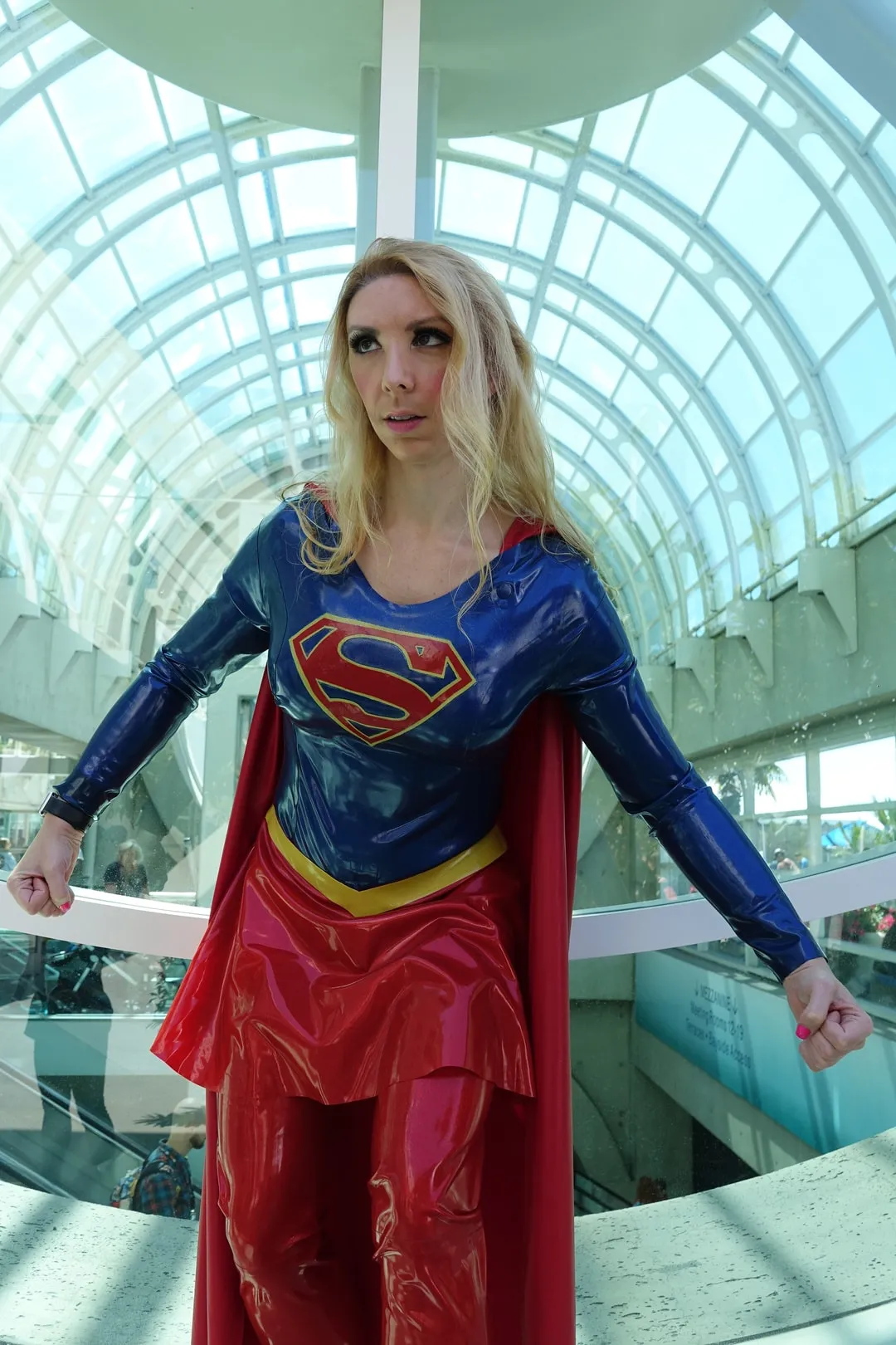 Jerikandra as Supergirl from the CW series in a blue and red latex suit with the S shield and cape, under a glass atrium roof.