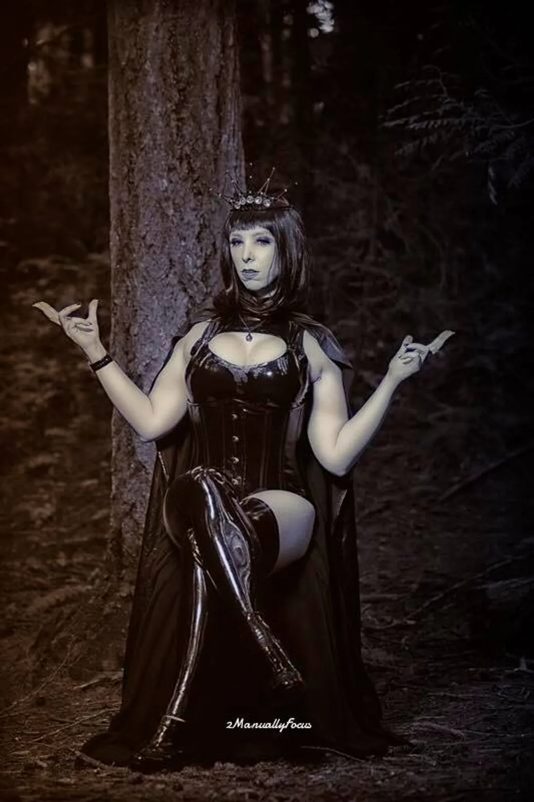 Jerikandra as Vampire Queen Andrastea in a black latex gown and crown with pale makeup, seated in a dark forest.
